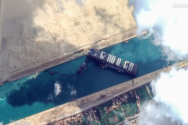 Esta imagem de satélite divulgada pela Maxar Technologies mostra rebocadores e dragas tentando libertar o MV Ever Given em 26 de março de 2021, no Canal de Suez. O navio porta-contêineres, com tamanho de mais de quatro campos de futebol, ficou paralisado diagonalmente por todo o canal, fechando a hidrovia nas duas direções. O bloqueio causou um enorme congestionamento de centenas de navios em cada extremidade do canal de 193 km (120 milhas) e grandes atrasos na entrega de petróleo e outros produtos