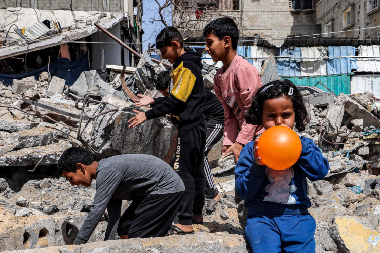 Uma menina sopra um balão enquanto seus meninos vasculham os escombros de um prédio destruído em Rafah, no sul da Faixa de Gaza, em 5 de abril de 2024, em meio ao conflito em curso no território palestino entre Israel e o grupo militante Hamas.
