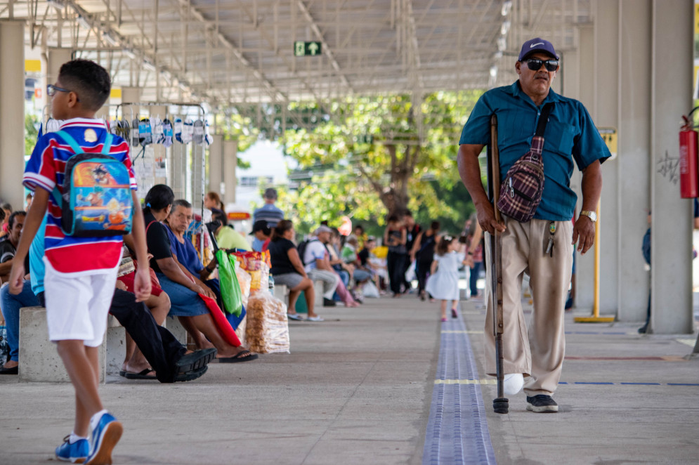 Passageiros circulam pelo terminal de ônibus do Papicu. Em primeiro plano, uma criança do sexo masculino com uma blusa do Fortaleza, com listras em vermelho azul e branco, vestindo também uma bermuda branca. Um pouco mais distante, um senhor de calça bege e blusa verde, portando uma muleta; (Foto: Samuel Setubal)