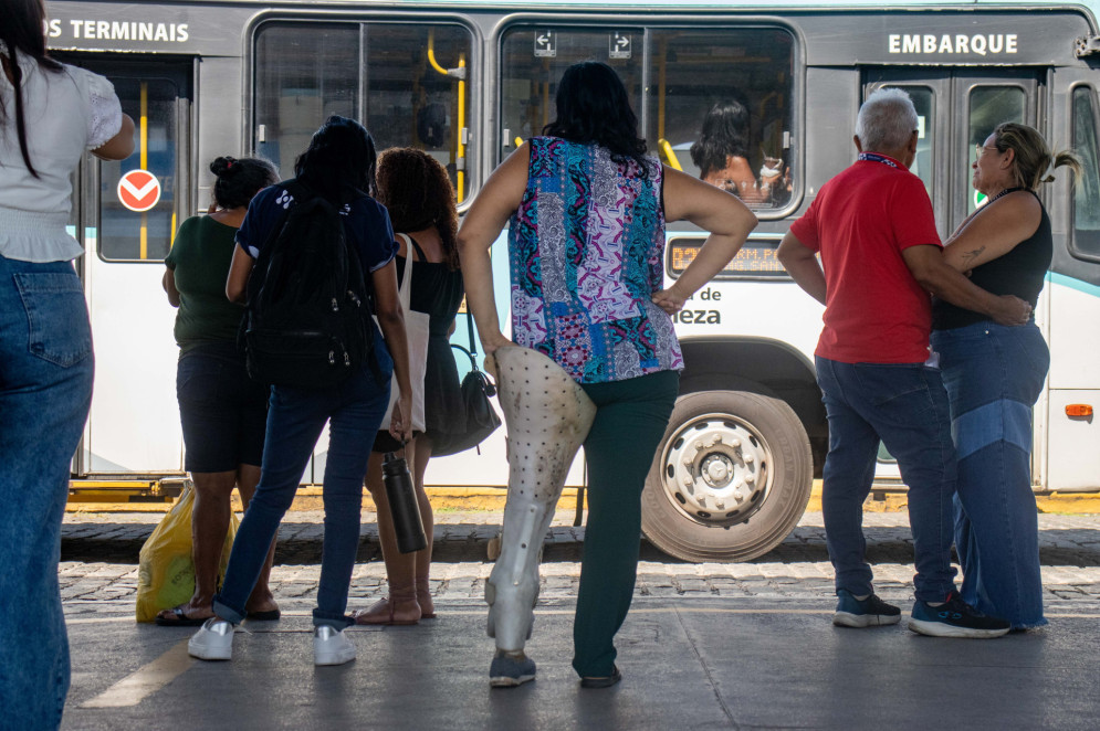 Passageira com prótese branca na perna esquerda espera ônibus em uma plataforma do terminal. A mulher veste uma blusa azul e branca, estampada com detalhes rosa, junto de uma calça verde escuro. Ela tem cabelos médios na altura do ombro e está com a mão direita na cintura. Ao fundo três mulheres e um casal também esperam o ônibûs. Em último plano, um ônibus azul com branco passa atrás de todos eles